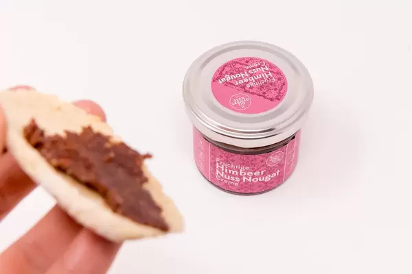 Fruchtige Himbeer Nuss Nougat Creme im Glas mit Aufstrich auf Brot im Vordergund
