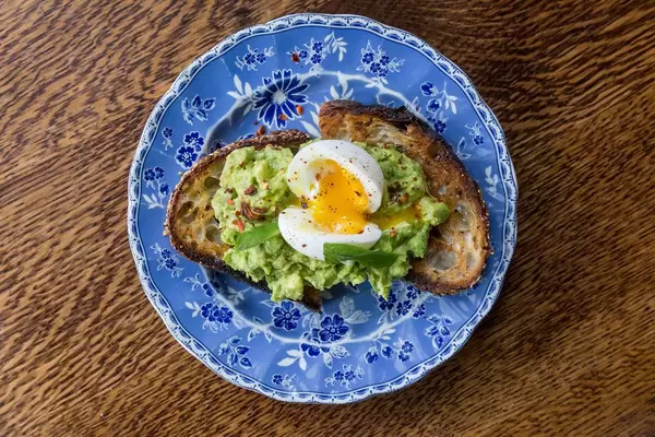 Frühstück mit Avocado, Ei, Basilikum, Limette, Chili und Mehrkornbrot auf einem blauen, blumendekorierten Teller, Aufnahme von Oben