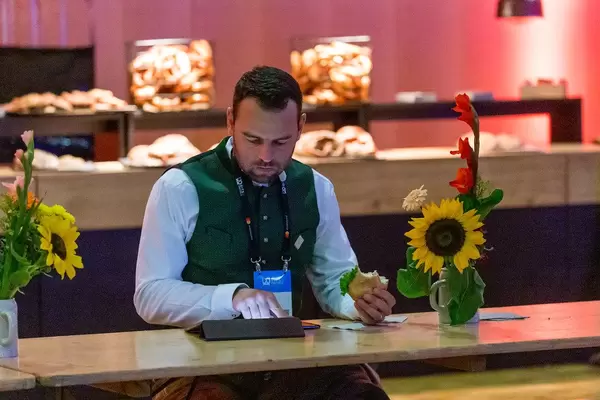 Frühstückspause auf der Bits & Pretzels Messe: Mann in bayrischer Tracht sitzt mit Brötchen an der Holzbar und nutzt das Tablet