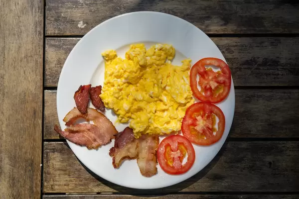 Frühstücksteller mit Rührei, gebratenen Speckstreifen und Tomatenscheiben von oben auf einem Holztisch fotografiert