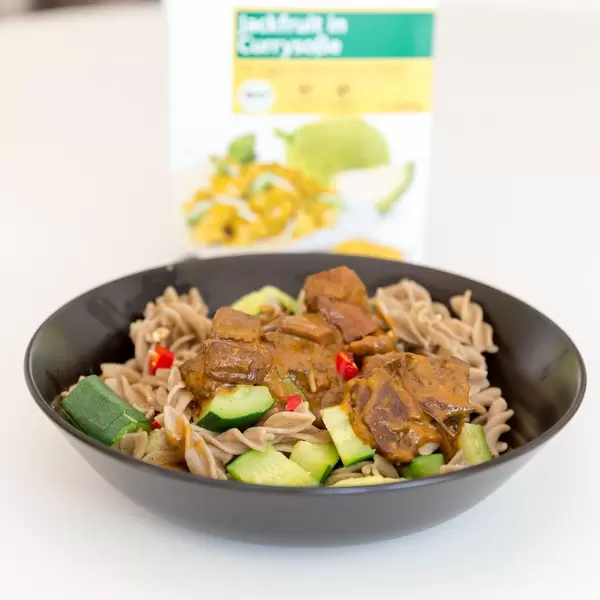 Fusilli mit Jackfruit, Gurkeln und Paprika in Currysauce mit dem Verpackung des veganen Instant-Gerichtes im Hintergrund
