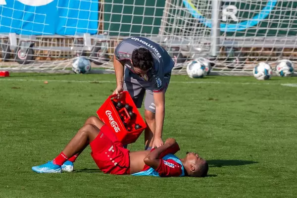 Fußballer Leon Bailey liegt erschöpft auf dem Rasen, Trainer kühlt ihn mit Coca Cola Flaschen