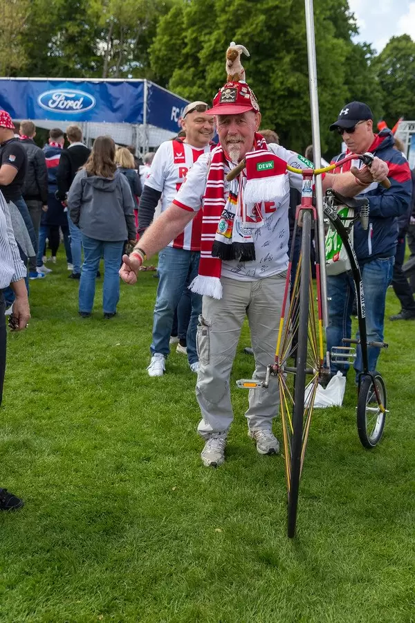Fußballfan mit 1. FC Köln Maskottchen auf verrücktem Hut, schiebt ein Hochrad in Farben der Deutschlandflagge im RheinEnergie- Stadion Park
