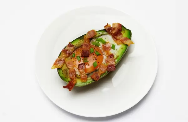 Gebackene halbe Avocado gefüllt mit Ei, Tomate und knusprigem Speck auf weißem Teller