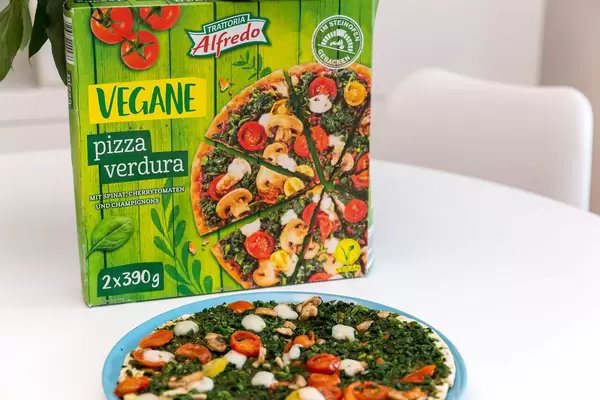 Gebackene Pizza Verdura von Trattoria Alfredo, mit Spinatbelag, Champignons und Cherrytomaten vor dem Pizzakarton auf einem weißen Tisch