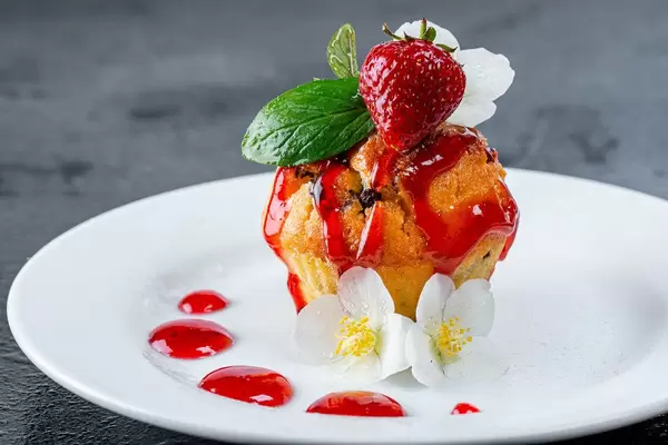 Gebackener Muffin mit frischer Erdbeere und Beerensauce als süßer Nachtisch, dekoriert mit weißen Blütenblättern
