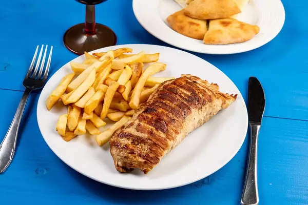 Gefülltes Schweinefleisch mit Pommes Frites angerichtet auf weißem Teller, mit Besteck auf blauem Holztisch