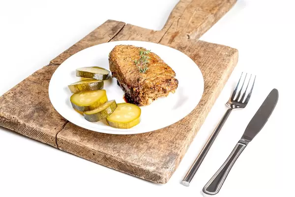 Gegrillter Kebab mit geschnittenen Gewürzgurken und Kräuter, neben Besteck auf einem rustikalen Holzbrettchen