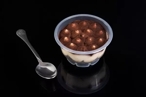 Gekauftes Tiramisu - Dessert im Plastikbecher, neben einem Löffel auf schwarzer, spiegelnder Oberfläche