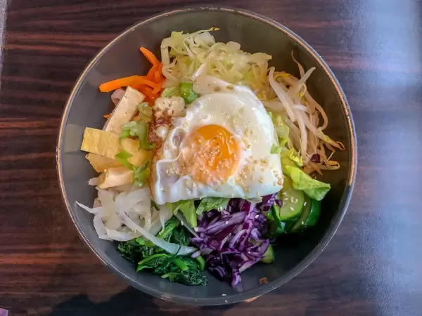 Gemischter Salat mit Tofu, Gurken, Sojasprossen, Karotten, Spiegelei und Sesam in Metallschüssel auf Holztisch