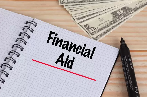 Geöffnetes Notebook mit rot unterstrichenem schwarzem Text Financial Aid mit schwarzem Filzstift und Dollar Geldscheinen auf einem Holztisch