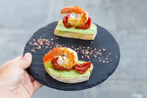 Geröstetes Brot, King Prawns, Avocado-Aufstrich, getrocknete Tomaten auf einem runden Brett bei Q11