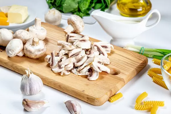 Geschnittene Champignons auf einem Küchenbrettchen, neben Pasta, Knoblauch und Lauchzwiebeln