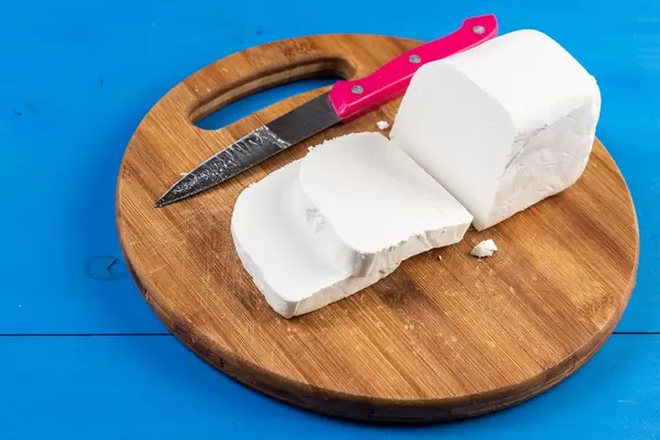 Geschnittener, weißer Käse mit Messer auf einem runden Holzteller, auf einem blauen Holztisch