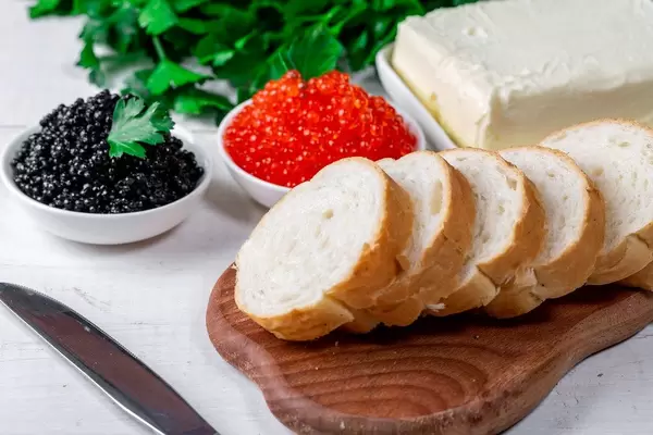 Geschnittenes Weißbrot neben Butter, schwarzem Kaviar und roten Lachskaviar als Delikatesse, auf einem weißen Holztisch
