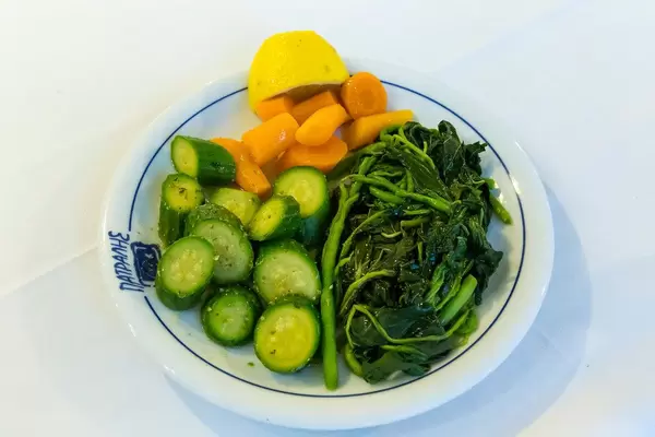 Gesunde Beilage zum Mittagessen, mit Blattspinat, Gurken, Möhren und einer halben Zitrone auf einem weißen Tisch