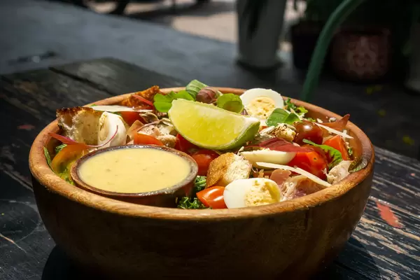 Gesunde Salat Bowl mit Hähnchen, gekochten Wachteleiern, Kirschtomaten, Parmesan Käse, Basilikum, Geröstetem Brot, Limette und Honig-Senf Sauce in einer Holzschüssel Nahaufnahme