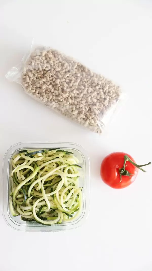 Gesunde Zucchini-Nudeln "Zoodles" neben einer Tomate und fleischloser, veganer Sonnenblumen-Bolognese, auf weißem Untergrund