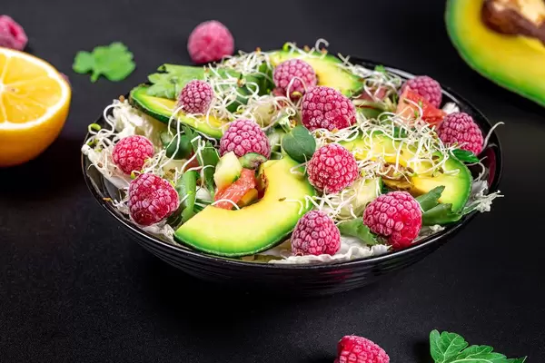 Gesunder Diät Salat mit grünem Salat, Avocado-Scheiben, Himbeeren, Mikrogrün und anderen Zutaten in einer schwarzen Schale auf schwarzem Hintergrund