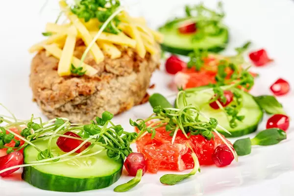 Gesundes Essen mit Kotelett, Käse, Gurkenscheiben, Granatapfelkernen, Pampelmuse und Mikrogrün auf einem weißem Teller Nahaufnahme