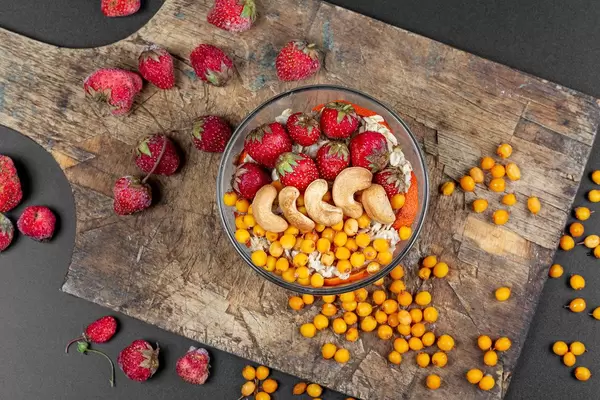 Gesundes Frühstück in einer Glasschale mit Haferflocken, Cashew Nüssen, Erdbeeren und Sanddornbeeren auf einem Holzschneidebrett von oben fotografiert
