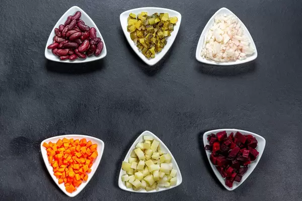 Gewürfelte Karotten, Kartoffelrüben, Bohnen, eingelegte Zwiebeln und Gurken in Dreiecksschalen auf schwarzem Hintergrund - Das Konzept des Kochens