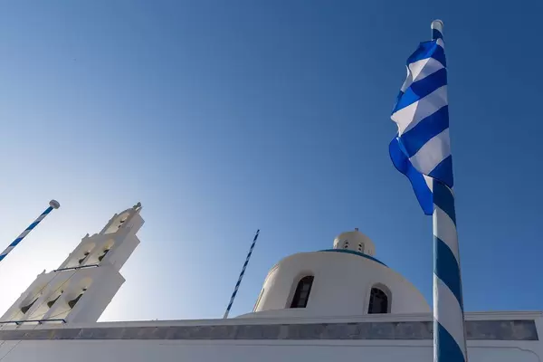Greek flag wrapped on a white and blue mast with white church bells and blue dome in the background