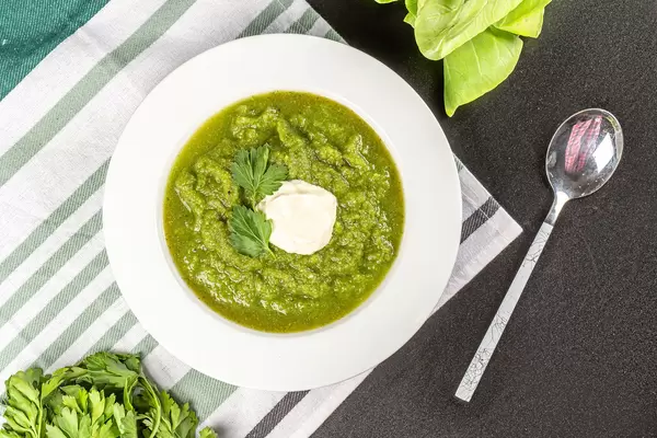 Green soup puree of spinach in white plate with a spoon on a dark background, top view