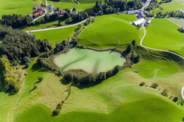 Grüne Hügel, Bäume und kleiner Frauensee mit hellgrünem Wasser bei Kramsach, Tirol. Luftbild