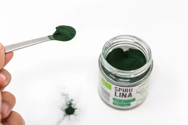 Grünes Spirulinapulver auf einem Löffel, neben dem Verpackungsglas auf einem weißen Tisch