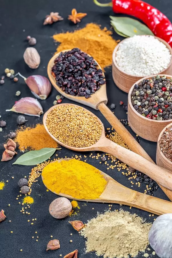 Gruppierung verschiedener Gewürze - Kurkumapulver, Senfkörner und Berberitzen auf Holzlöffeln mit Sesam, buntem Pfeffer und Leinsamen in Holzschälchen, Knoblauchzehen, Sternanis und  Piment auf schwarzem Hintergrund