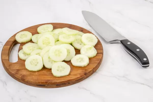 Gurkenscheiben als gesunde Salatbeigabe, liegen neben einem Küchenmesser auf einem runden Küchenbrettchen aus Holz