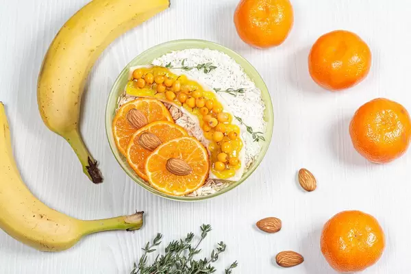 Haferflocken mit Mandarinenscheiben, Bananen, Sanddornbeeren, und Kokosraspeln auf einem weißen Holztisch