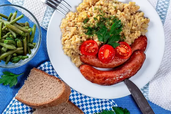 Haferflocken-Porridge mit Bratwürstchen, Tomatenscheiben und rustikalem Brot, auf einem blau-weiß gedeckten Tisch, im Bayrischen Look