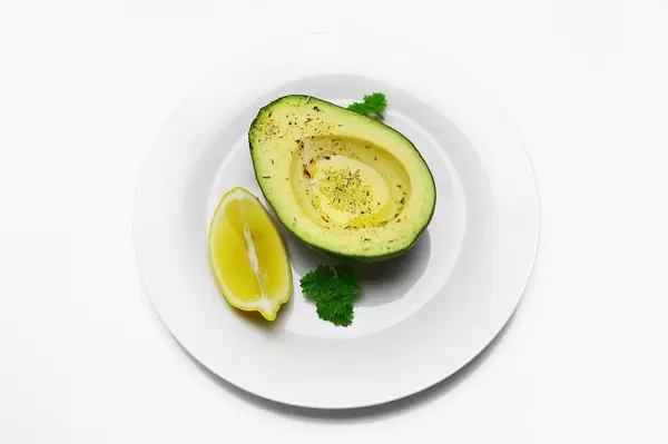Halbierte und entkernte Avocado mit Kräutern bestreut, neben Zitrone und Petersilie auf Teller