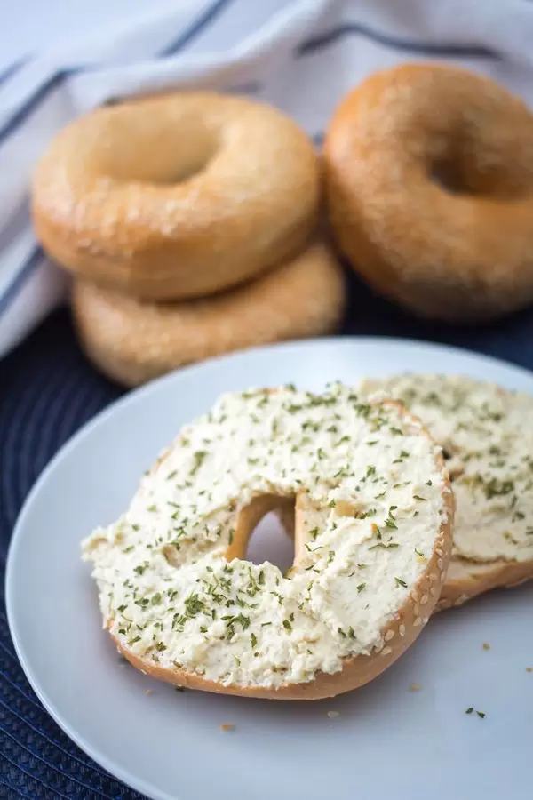 Halbierter Bagel, bestrichen mit Frischkäse und Kräutern, im Hintergrund weitere Bagels
