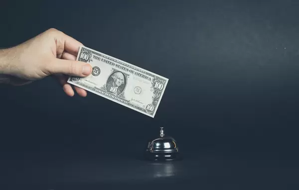 Hand hält Banknote über silberne Serviceklingel vor dunklem Hintergrund