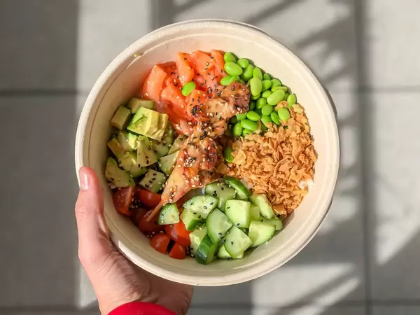 Hand hält das Tagesgericht Surf and Turf Bowl von Eat Simple, mit Hähnchen, Röstzwiebeln, Favabohnen und Gemüse