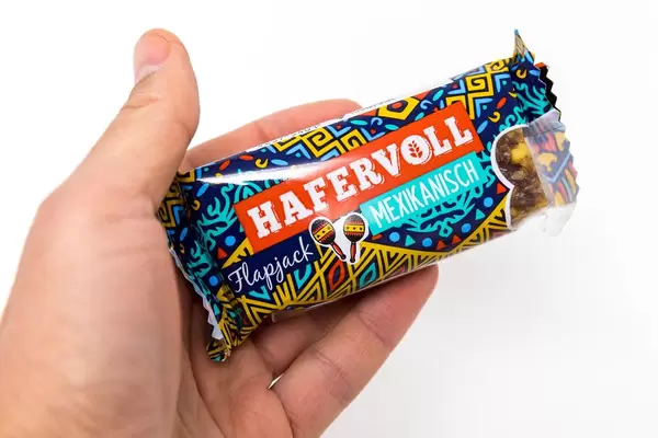 Hand hält den Hafervoll Flapjack Mexikanisch - Müsliriegen in bunter Verpackung mit zwei Maracas aufgedruckt, vor weißem Hintergrund