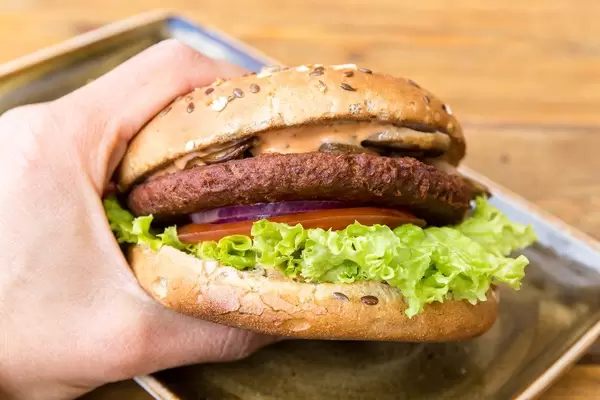 Hand hält den veganen, fleischlosen Burger "Naturbursche" der Marke Moving Mountains, mit karamellisierten Zwiebeln, Champions und Salat