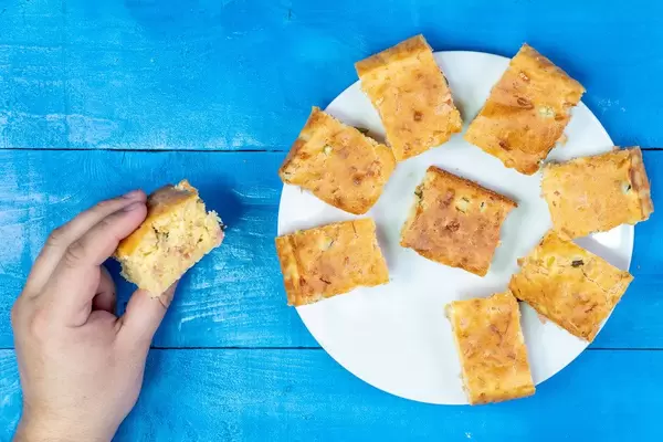 Hand hält ein hausgemachtes Kuchenstück, gefüllt mit Käse und Schinken, auf einem blauen Tisch