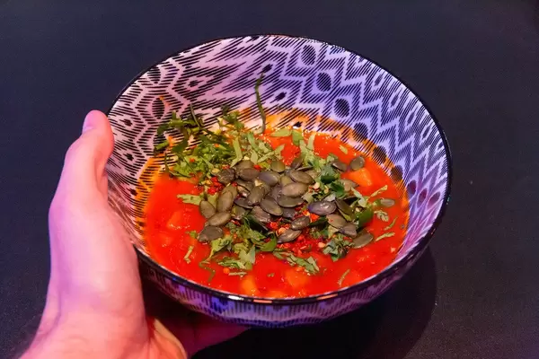 Hand hält eine schwarz-weiße Schüssel mit Tomaten-Sellerie-Mohrensuppe mit Samen und frischen Kräutern