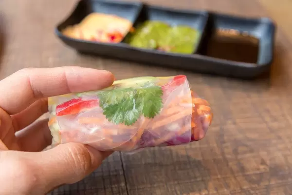 Hand hält eine Sommerrolle gefüllt mit Avocado, Mango, Paprika, Rotkohl, Karotten und Reisnudeln mit verschiedenen Saucen im Hintergrund