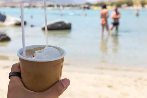 Hand hält einen kalten, griechischen Frappé mit Eiskugel, am Strand von Paros