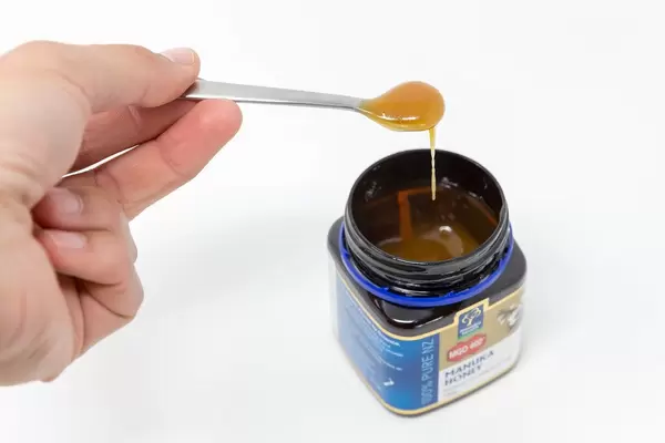 Hand hält einen Löffel mit flüssigem Honig über da Honigglas "Manuka Honey"