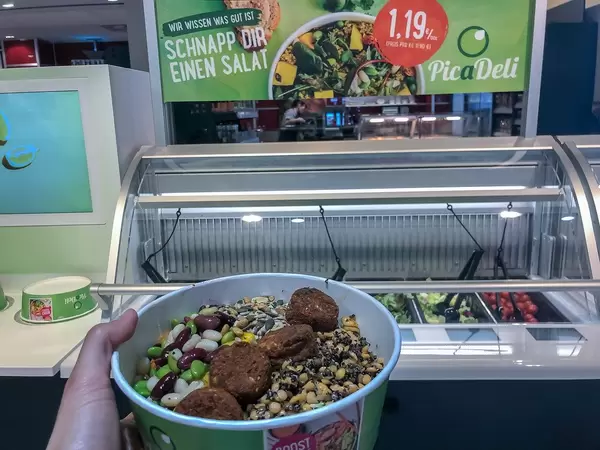 Hand hält einen Salatmix in der grünen To-Go-Salatschüssel, vor der Angebotstafel der digitalen PicaDeli Salatbar