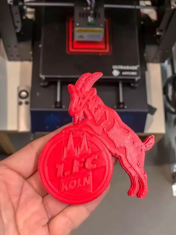 Hand hält Geißbock Hennes, das Maskottchen des 1. FC Köln als roter 3D-Druck vor dem 3D-Drucker
