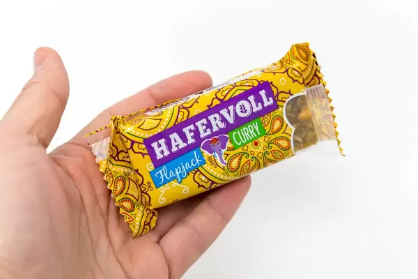 Hand hält Hafervoll Flapjack Curry Riegel in Verpackung im indischem Look vor weißem Hintergrund, enthält Sojabohnen & Mangostücken