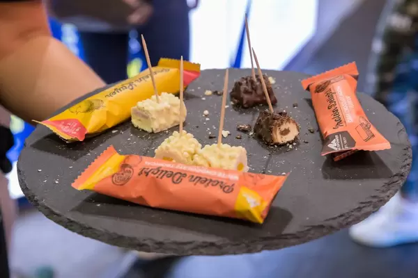 Hand hält Multipower Protein Delight Schokoladenbarren auf einen Probierteller in verschiedenen Geschmäcken: Zartbitterschokolade Moccha, Weiße Schokolade Tropical & Weiße Schokolade Erdbeere