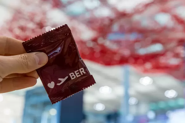 Hand hält Werbegeschenk mit Herz und BER-Logo vor dem roten "fliegendem Teppich" von Pae White